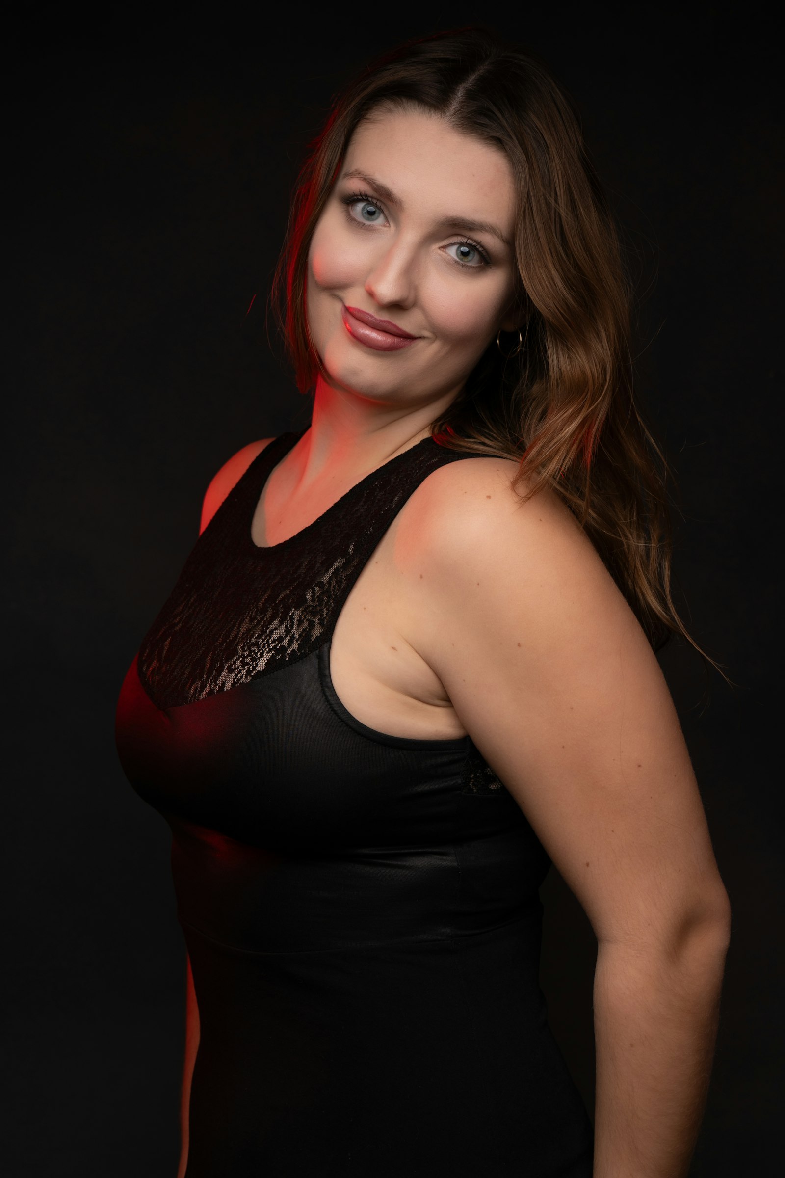 Woman with long wavy brown hair falling over one shoulder, in a black top against a dark studio background