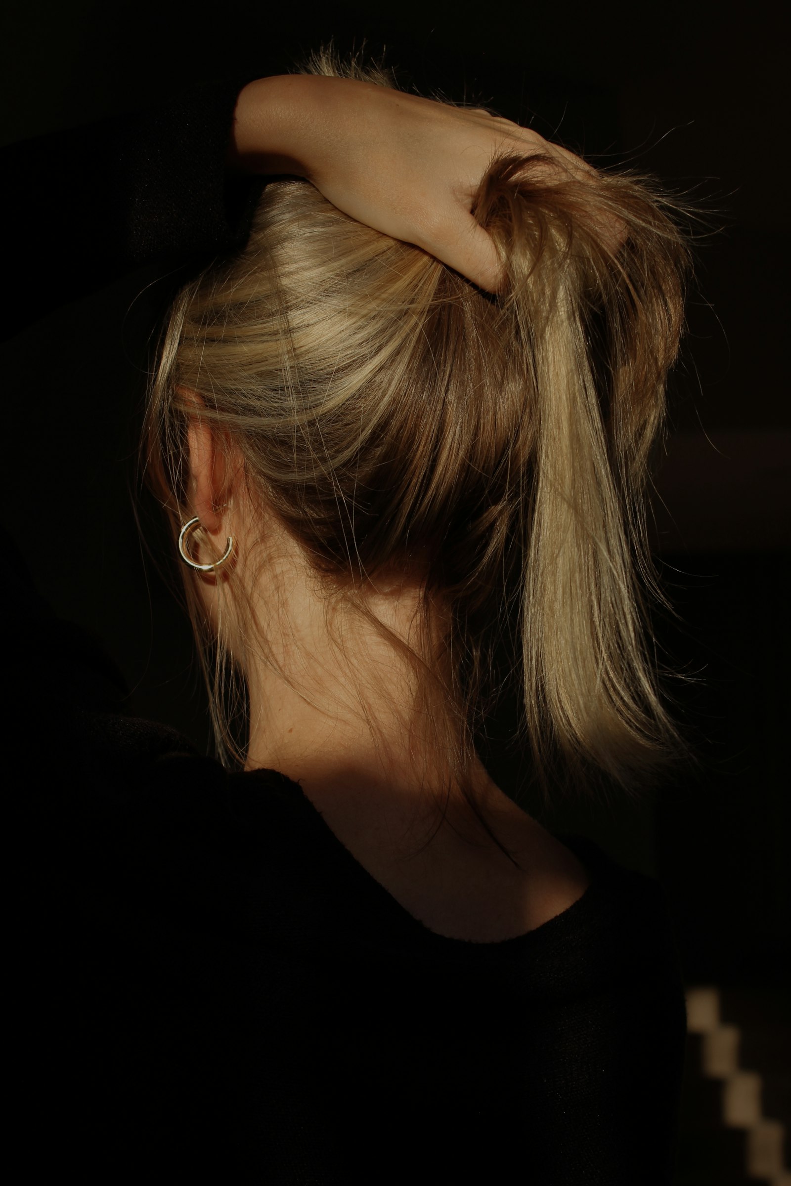 Back view of a woman gathering her blonde hair into a high ponytail with one hand