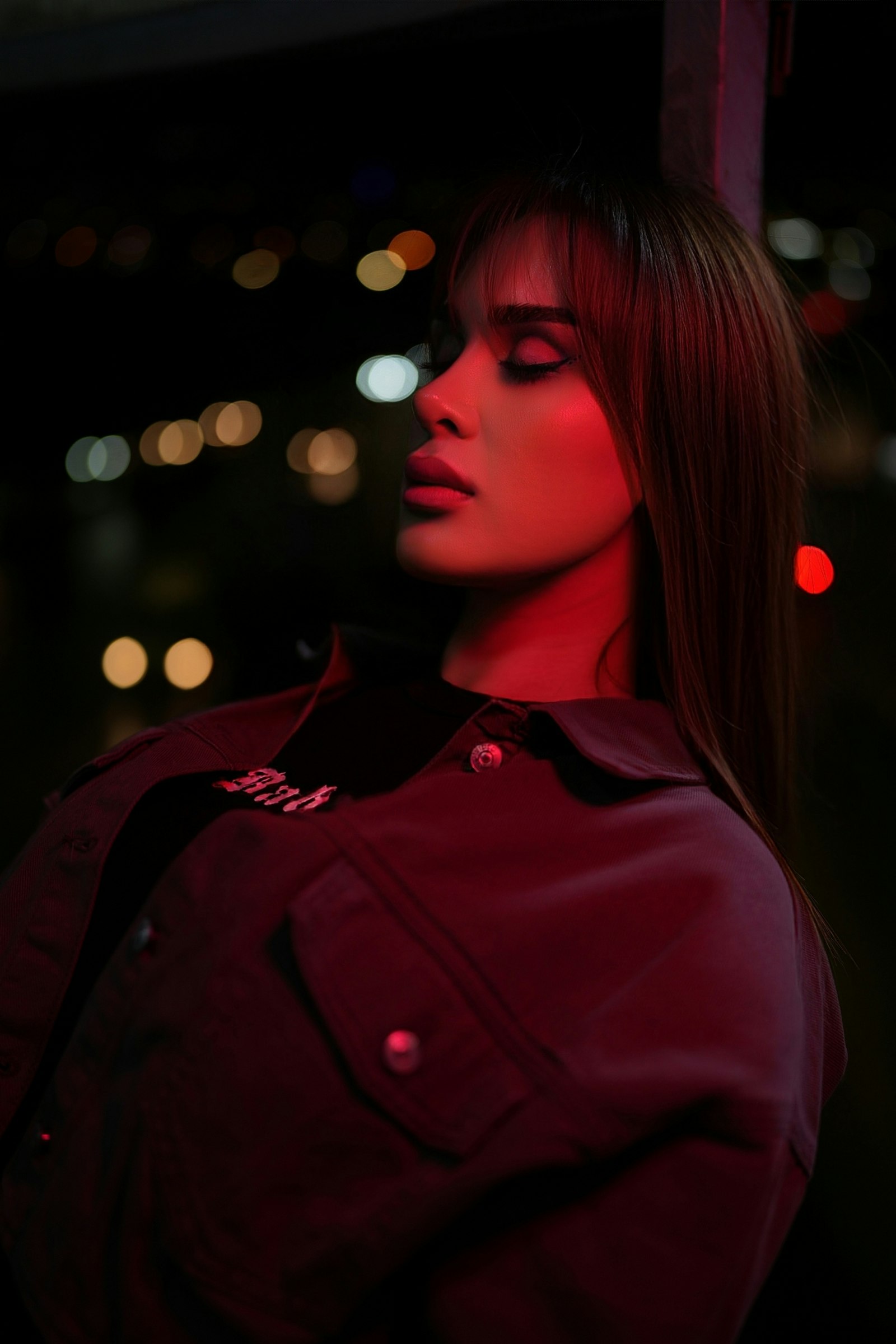 Woman in profile with long dark auburn hair and a fringe, lit red against out-of-focus city lights at night
