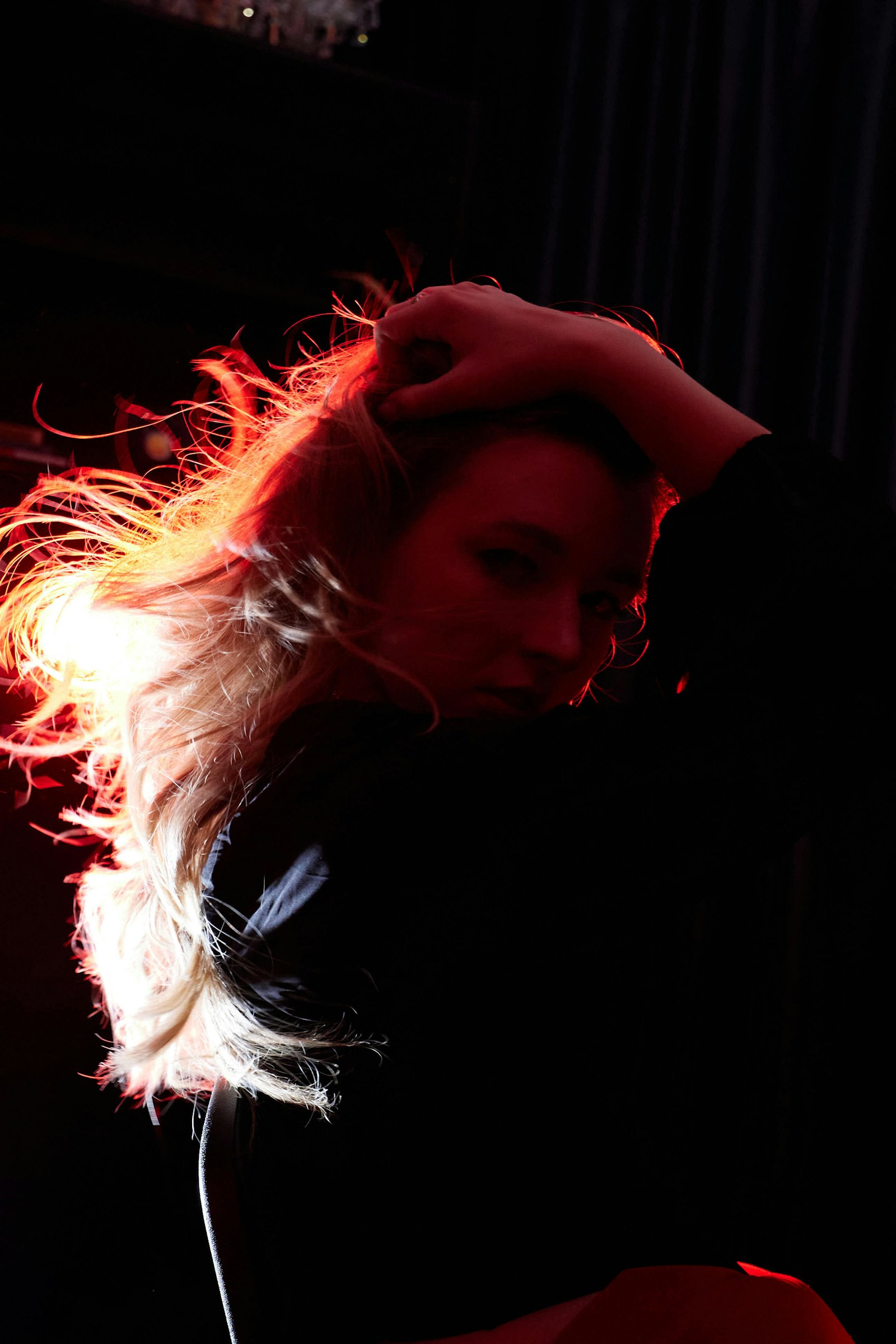 Woman with long blonde hair lit by strong red light against a black background, one hand pushed back through her hair